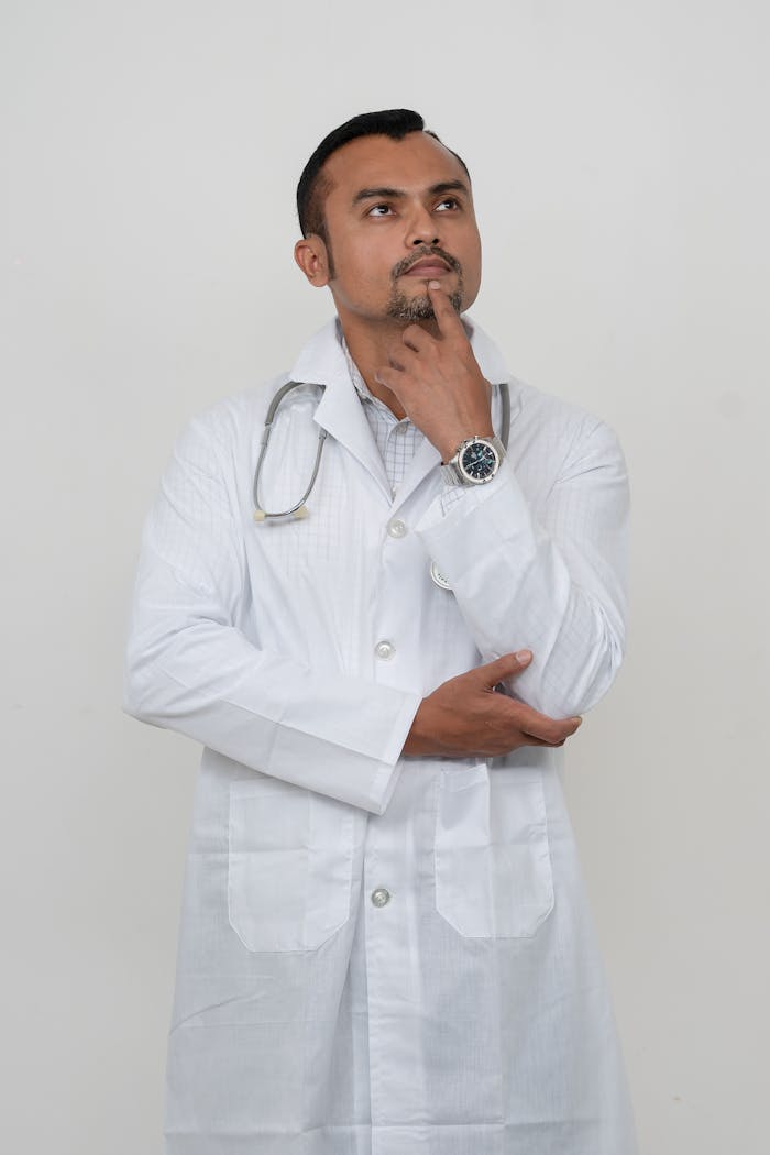 A thoughtful male doctor in a white coat, in a studio setting, posing with his hand on his chin.
