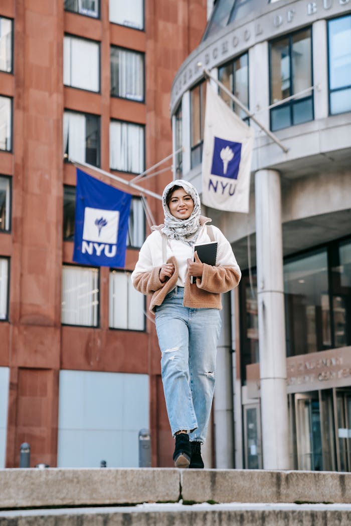 Cheerful student walking by a university building with NYU flags, embracing a diverse and urban education setting.