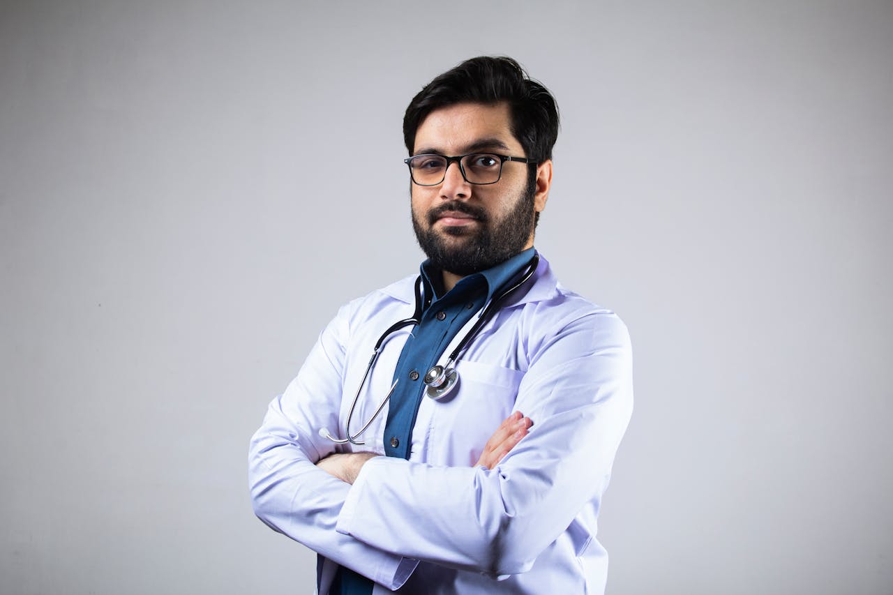 Portrait of a serious male doctor in a white coat with a stethoscope, arms crossed, studio background.
