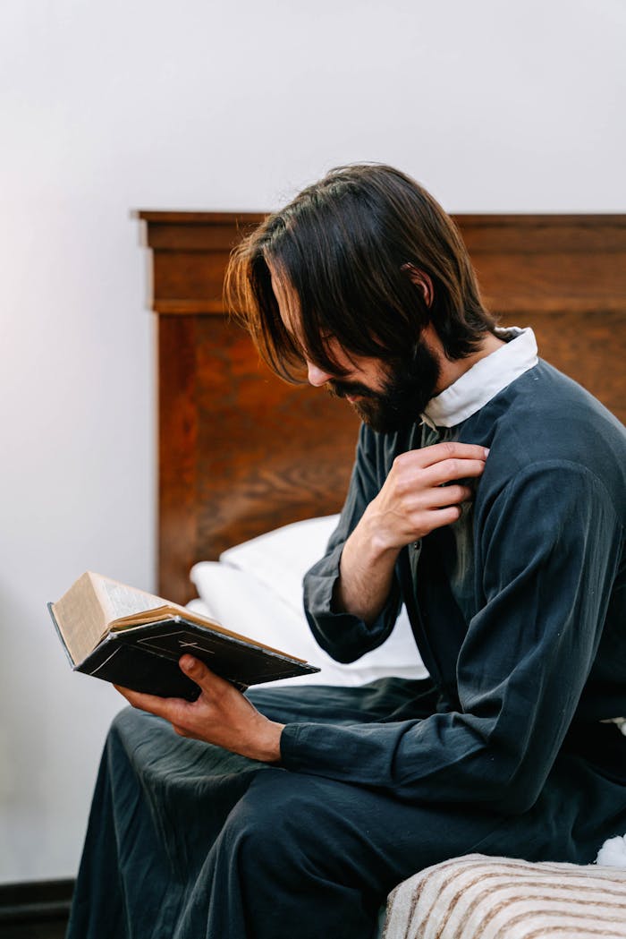 A seminarian in a robe sits on a bed, holding and reading a Bible, making the sign of the cross.